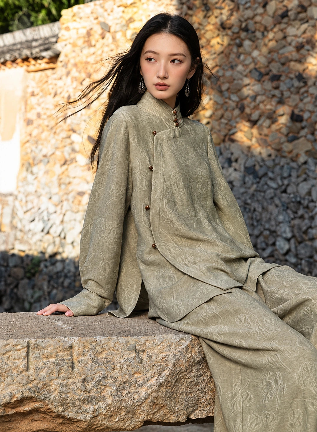 Model seated on a stone ledge wearing an antique sage jacquard shirt and matching trousers, showing the overlapping front panel.