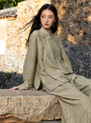 Model seated on a stone ledge wearing an antique sage jacquard shirt and matching trousers, showing the overlapping front panel.