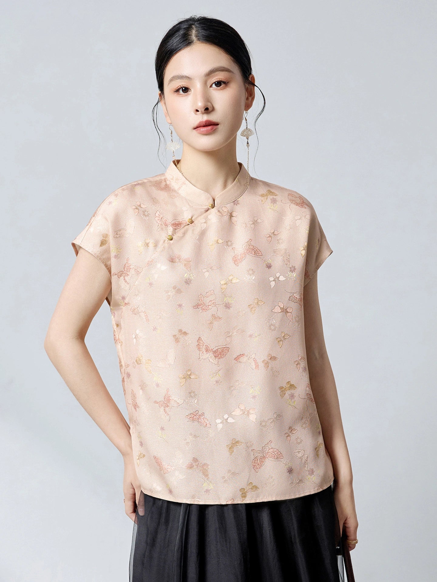 Model wearing an apricot 94% mulberry silk top with a stand collar and butterfly jacquard pattern, styled with a black skirt, featuring traditional frog buttons.