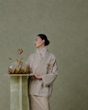 Model wearing an embroidered beige Chinese-inspired jacket beside a golden glass vase against a muted green wall.