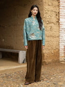 Full-length outfit featuring the Baishi teal jacquard jacket with brown wide-leg trousers beside a textured wall.