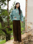Model wearing the Baishi teal jacket and brown trousers outdoors beside trees and a brick-edged wall.