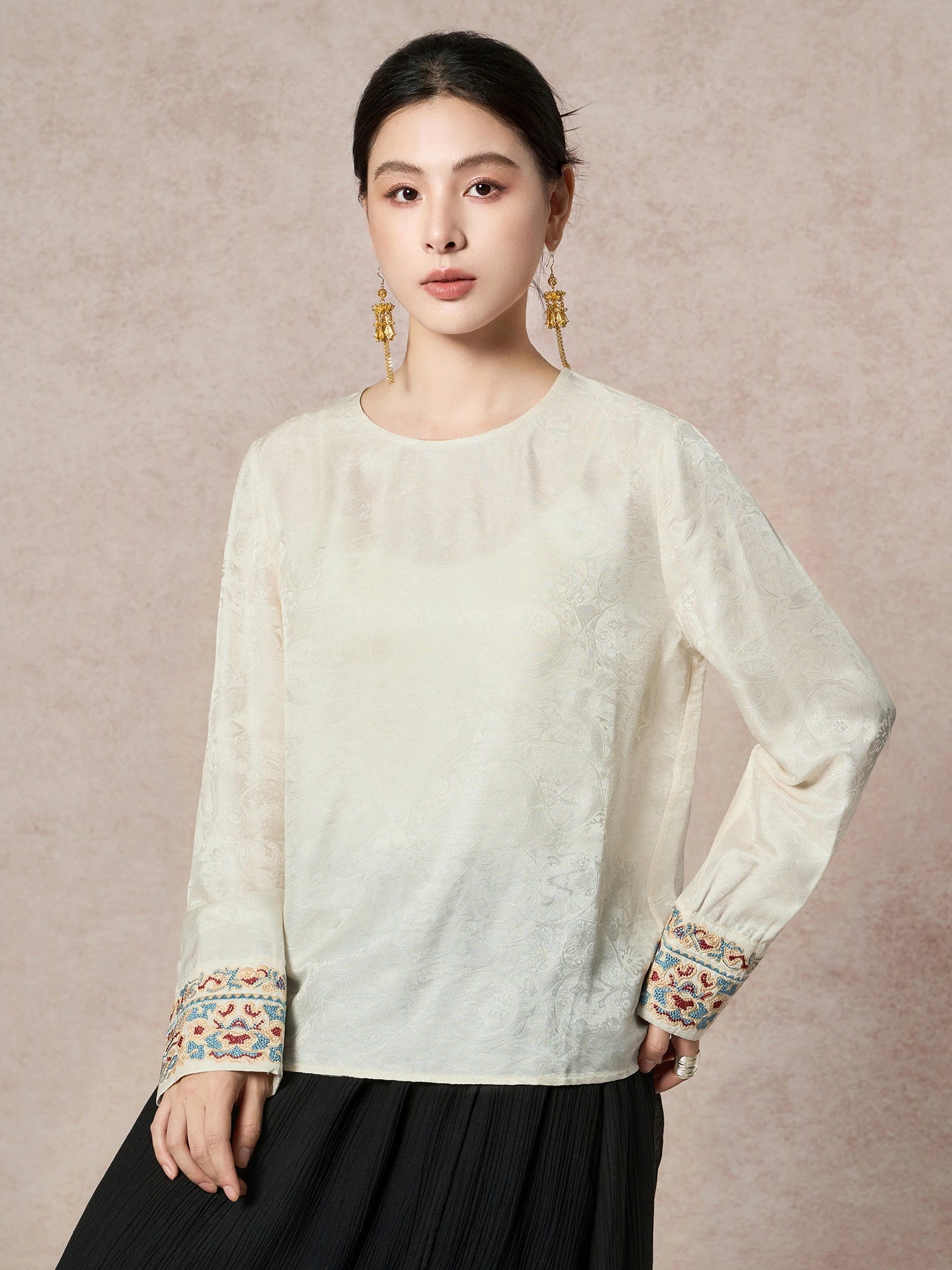 Model wearing a cream white long-sleeve blouse with a round neck and embroidered cuffs, paired with a black pleated skirt, showcasing the elegant silhouette.