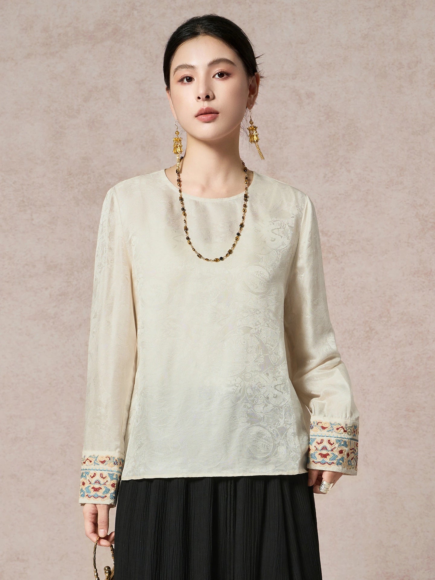Front view of a cream white silk-blend blouse with jacquard texture and bead embroidery on the cuffs, styled with a long beaded necklace.