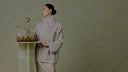 Model wearing a beige embroidered Chinese-inspired jacket beside a golden glass vase against a muted green textured wall.