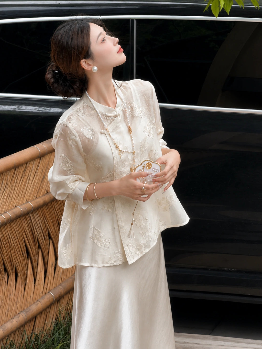 Model in a beige embroidered Cheongsam blouse and satin skirt, holding a fan and looking up, showcasing the relaxed fit and delicate embroidery.