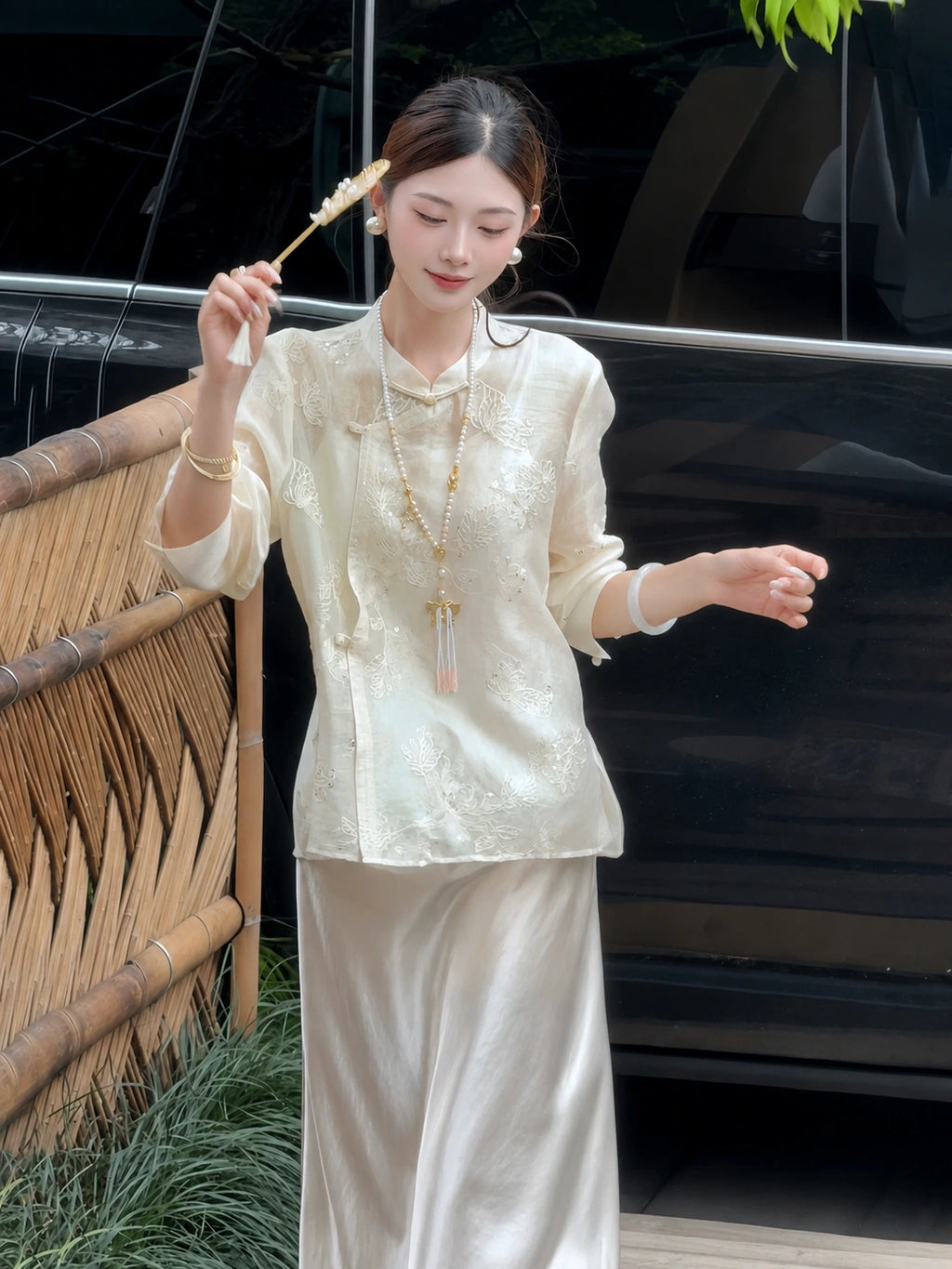 Full body shot of a beige embroidered Cheongsam blouse and matching satin skirt, styled with a pearl necklace and fan, crafted from 100% polyamide.