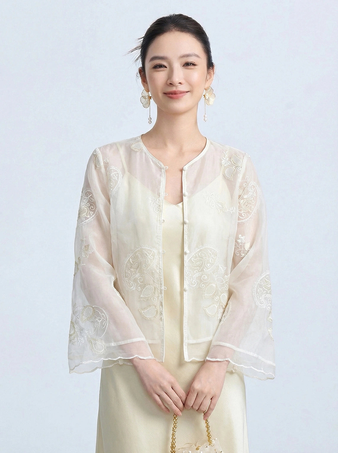 Full body shot of a cream Tencel embroidered cardigan with a front button closure and sheer fabric, styled with a gold handbag and cream mules.
