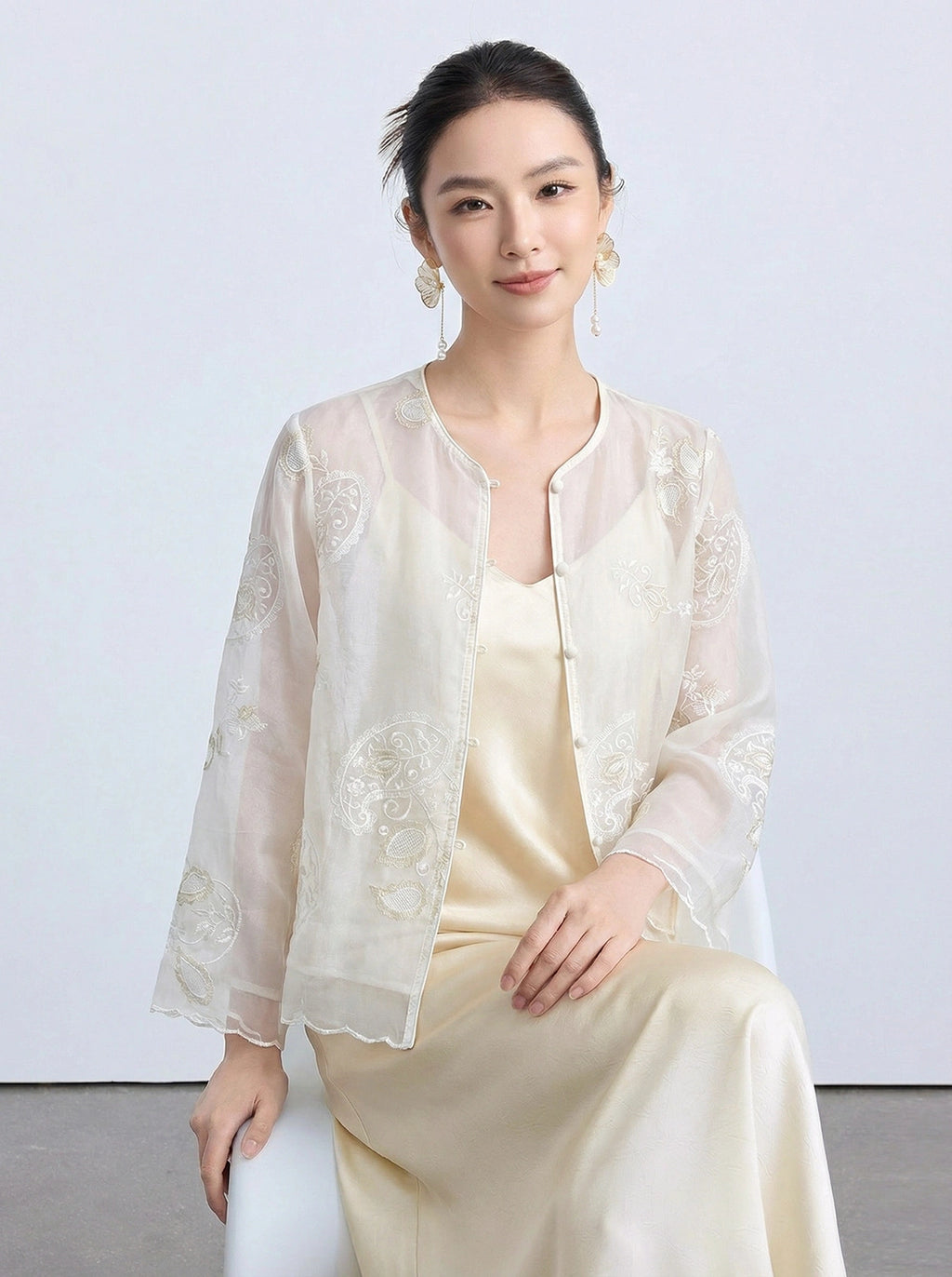 Model seated in a cream sheer Tencel cardigan featuring delicate embroidery and flared sleeves, paired with a satin dress.