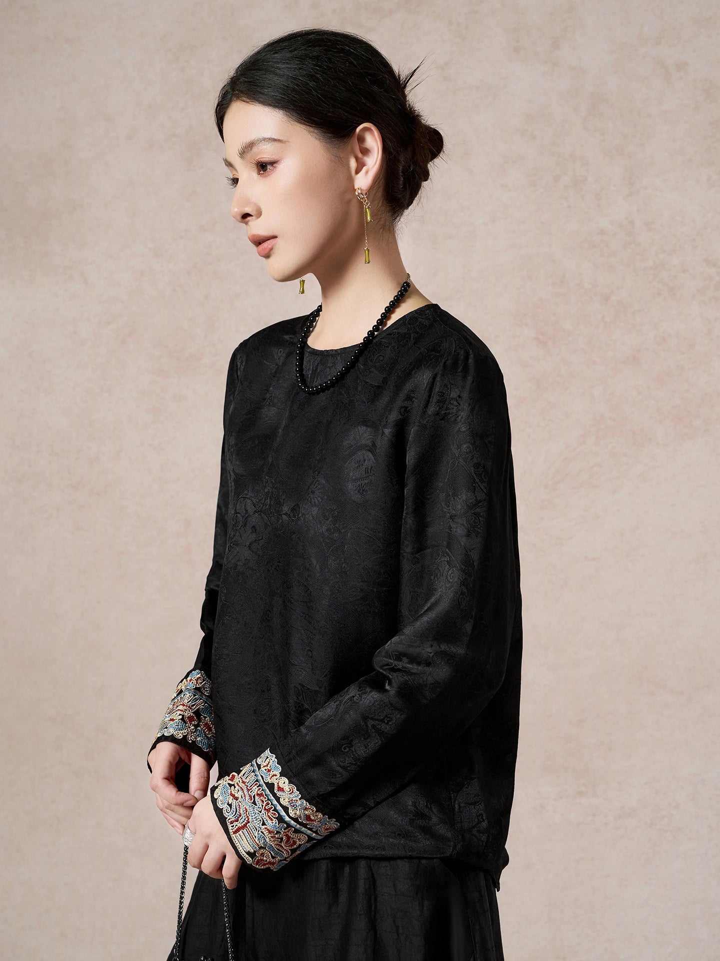 Model wearing a black long-sleeve silk-blend blouse with a round neck and intricate bead embroidery on the cuffs, showcasing the luxurious drape and detailing.