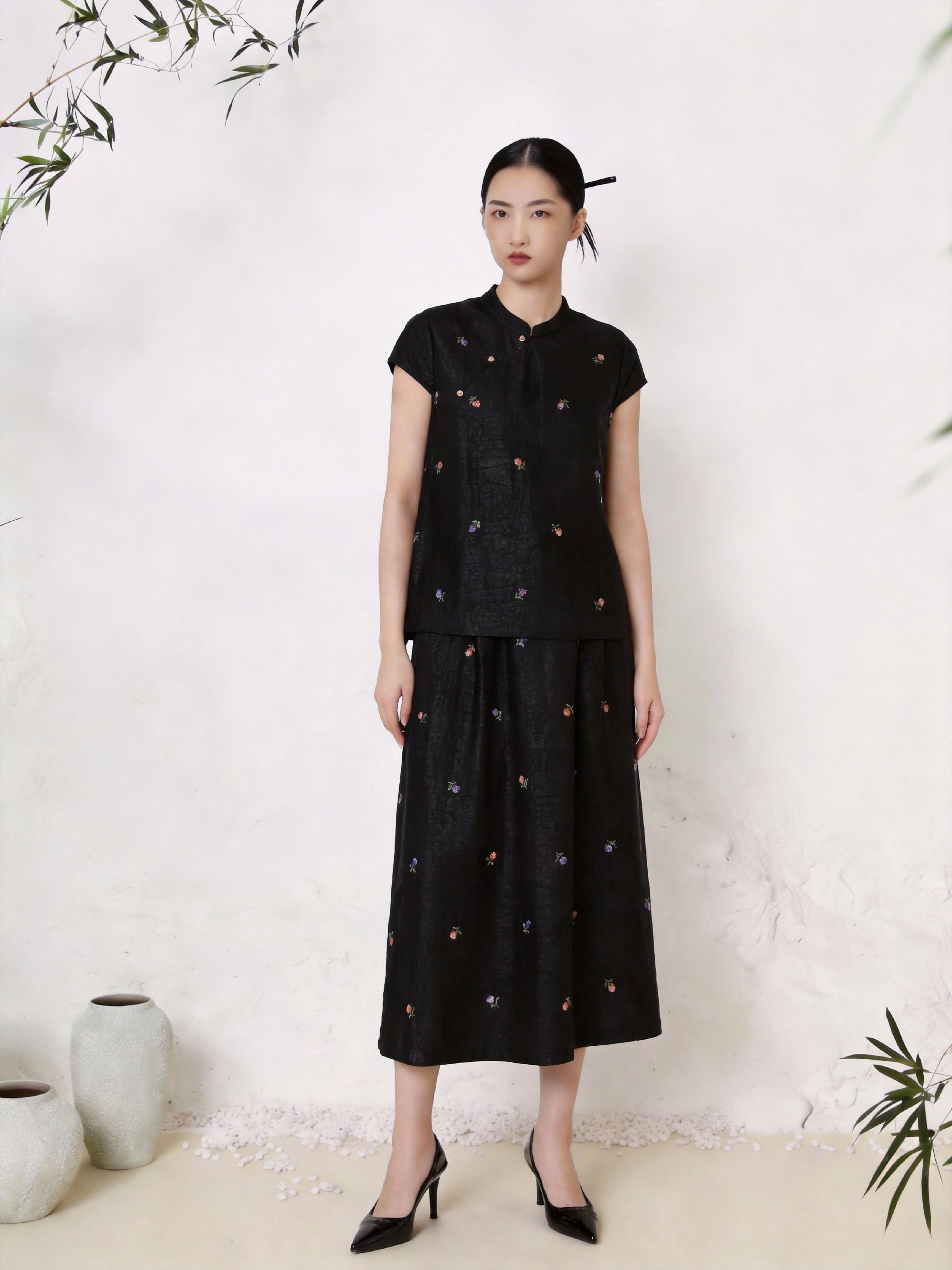 Full body shot of a black silk co-ord set featuring a short sleeve Cheongsam top with a mandarin collar and scattered colorful floral embroidery, paired with a matching A-line midi skirt with similar floral details, crafted from 100% mulberry silk.