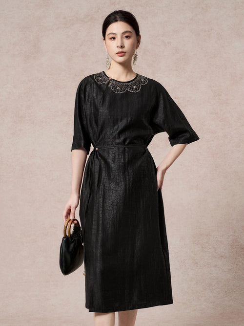 Front view of a black silk dress featuring intricate scalloped embroidery on the chest, a round neckline, and a side tie waist detail.