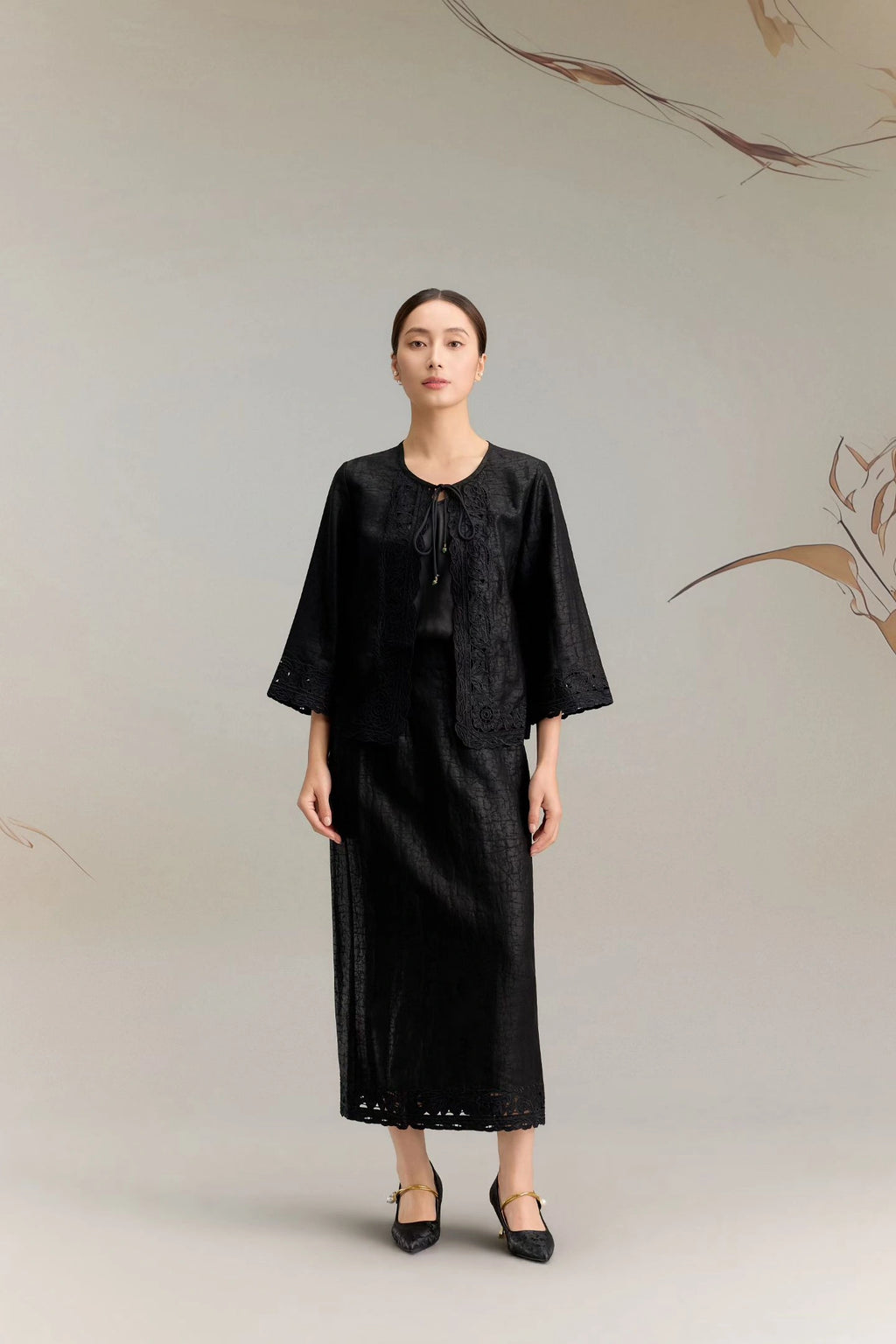 Full body shot of a black silk co-ord set featuring a mid-sleeve jacket top with delicate lace embroidery along the edges and a tie-front closure, paired with a matching A-line midi skirt with lace detailing at the hem, crafted from 100% mulberry silk.