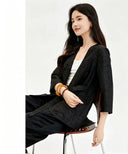 Side view of a black silk wrap blouse with three-quarter sleeves, V-neckline, and a tie waist, crafted from 100% mulberry silk, featuring a subtle jacquard texture, paired with white inner top and black pants.