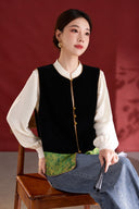 Seated model wearing the black velvet vest with green jacquard trim, an ivory blouse and jeans.