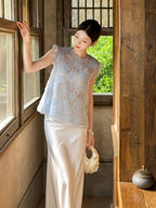 Model wearing a light blue floral embroidered top with sequins and ruffle sleeves, holding a white bag, styled for a refined New Chinese look.