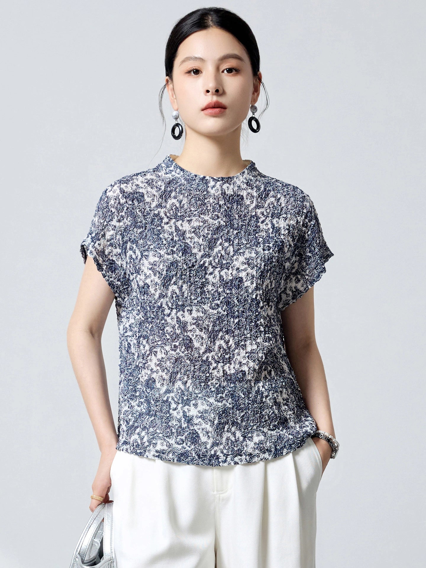 Front view of a women's blue and white printed silk blouse with short sleeves and a relaxed fit, paired with white trousers.
