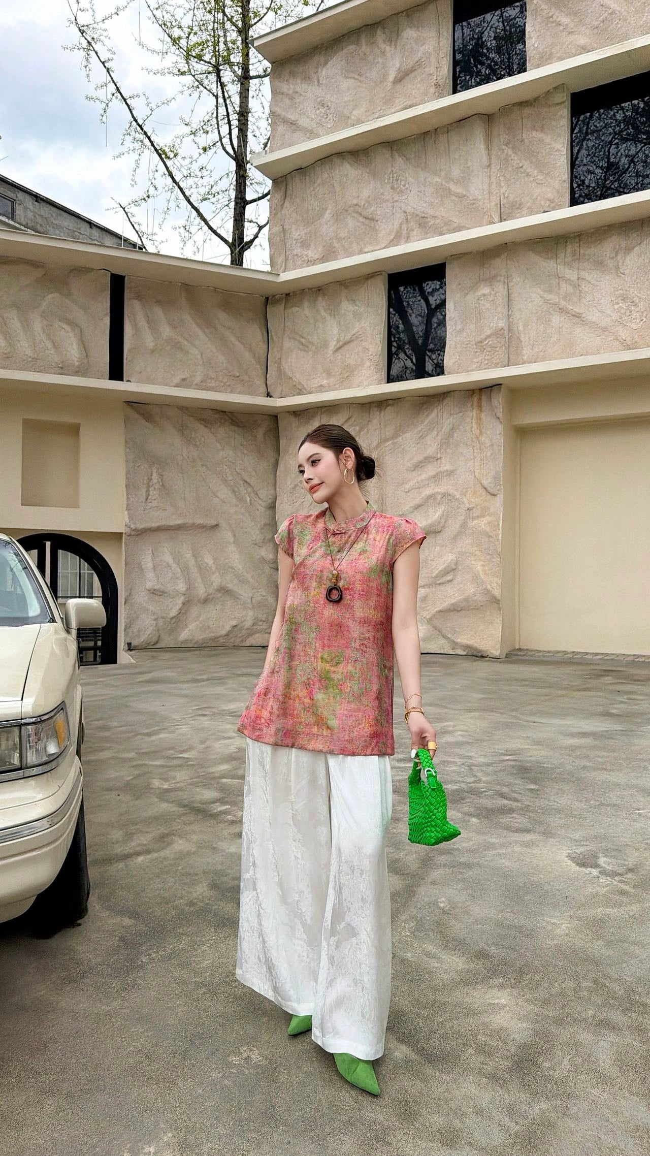 Front view of a women's pink Qipao top with traditional button detail and a watercolor floral jacquard texture, styled with white trousers and green heels.