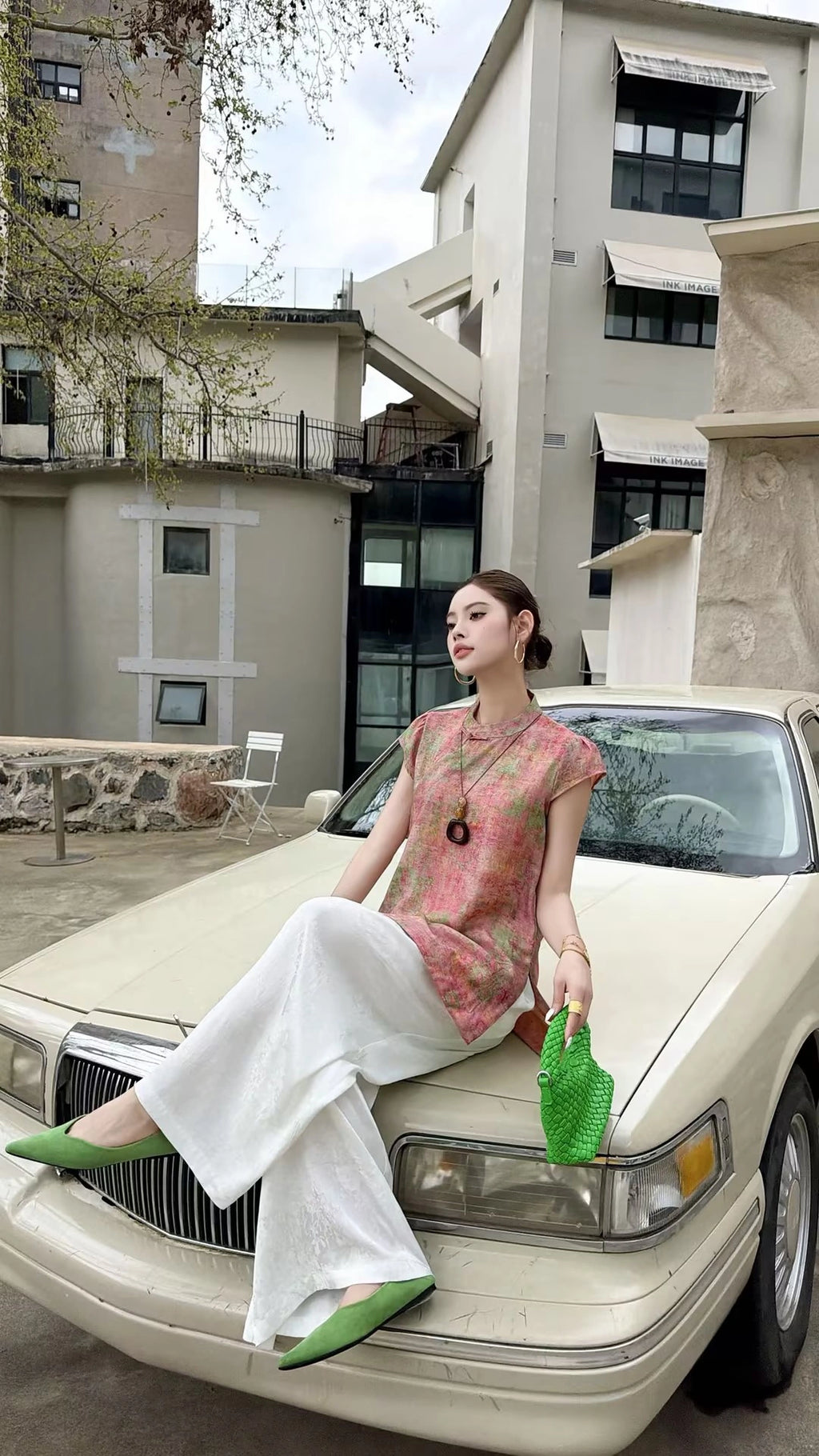 Sitting woman in a blush pink Cheongsam top with a mandarin collar, styled with white wide-leg pants and green heels, perfect for elegant summer styling.