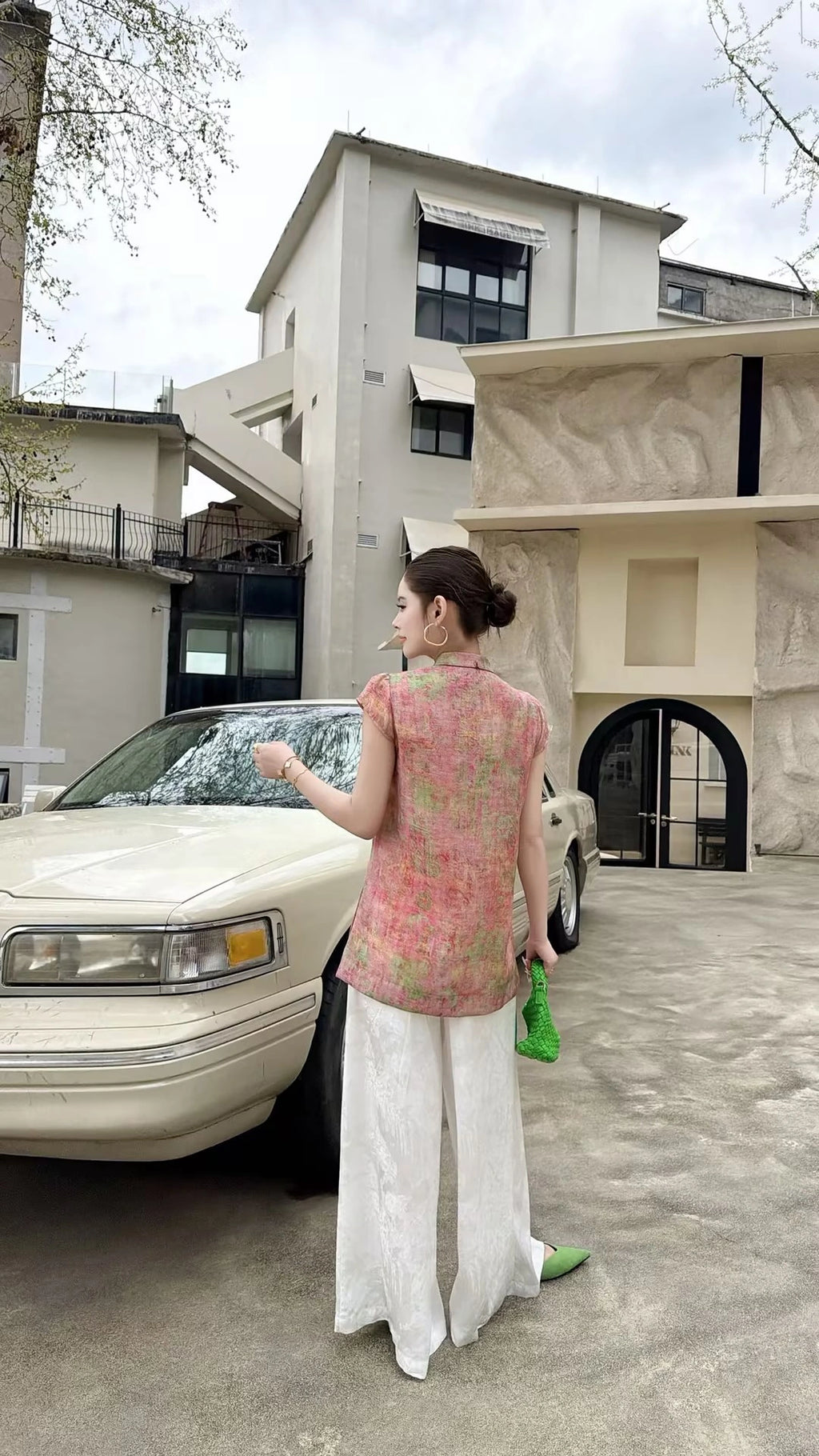 Back view of a blush pink Cheongsam top with cap sleeves and mandarin collar, crafted from 80% viscose and 20% nylon, paired with white wide-leg pants and a green bag.