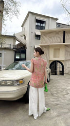 Back view of a blush pink Cheongsam top with cap sleeves and mandarin collar, crafted from 80% viscose and 20% nylon, paired with white wide-leg pants and a green bag.