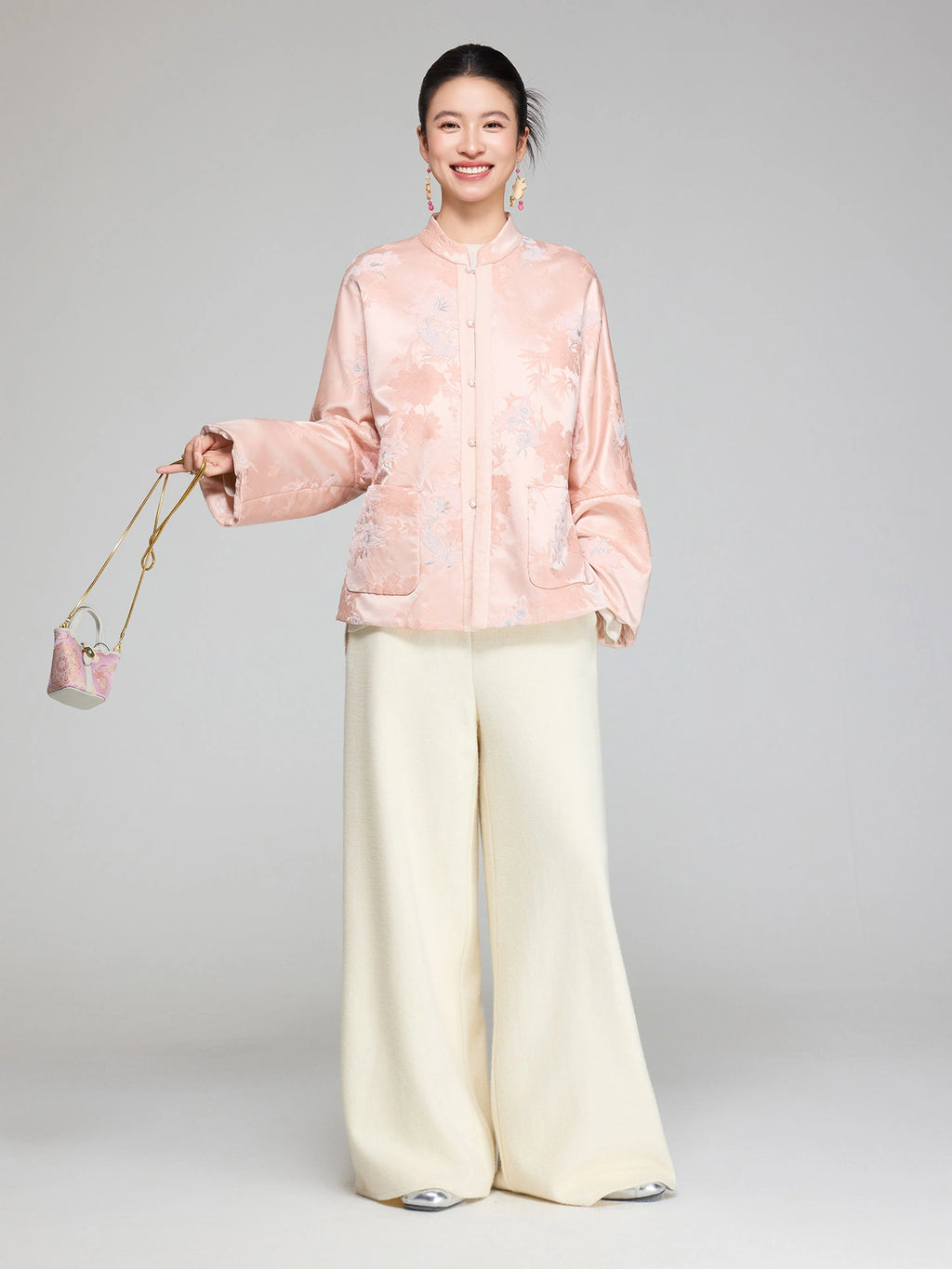 Woman wearing a blush pink floral jacquard padded jacket with pearl-style buttons and cream wide-leg trousers
