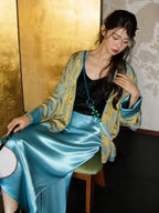 Seated styling image of a bright blue and gold V-neck jacket paired with a glossy water-blue skirt for an elegant commuter look.