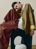 Seated model wearing a bronze mulberry silk-blend Chinese jacket with knotted buttons, golden cuffs and patch pockets.