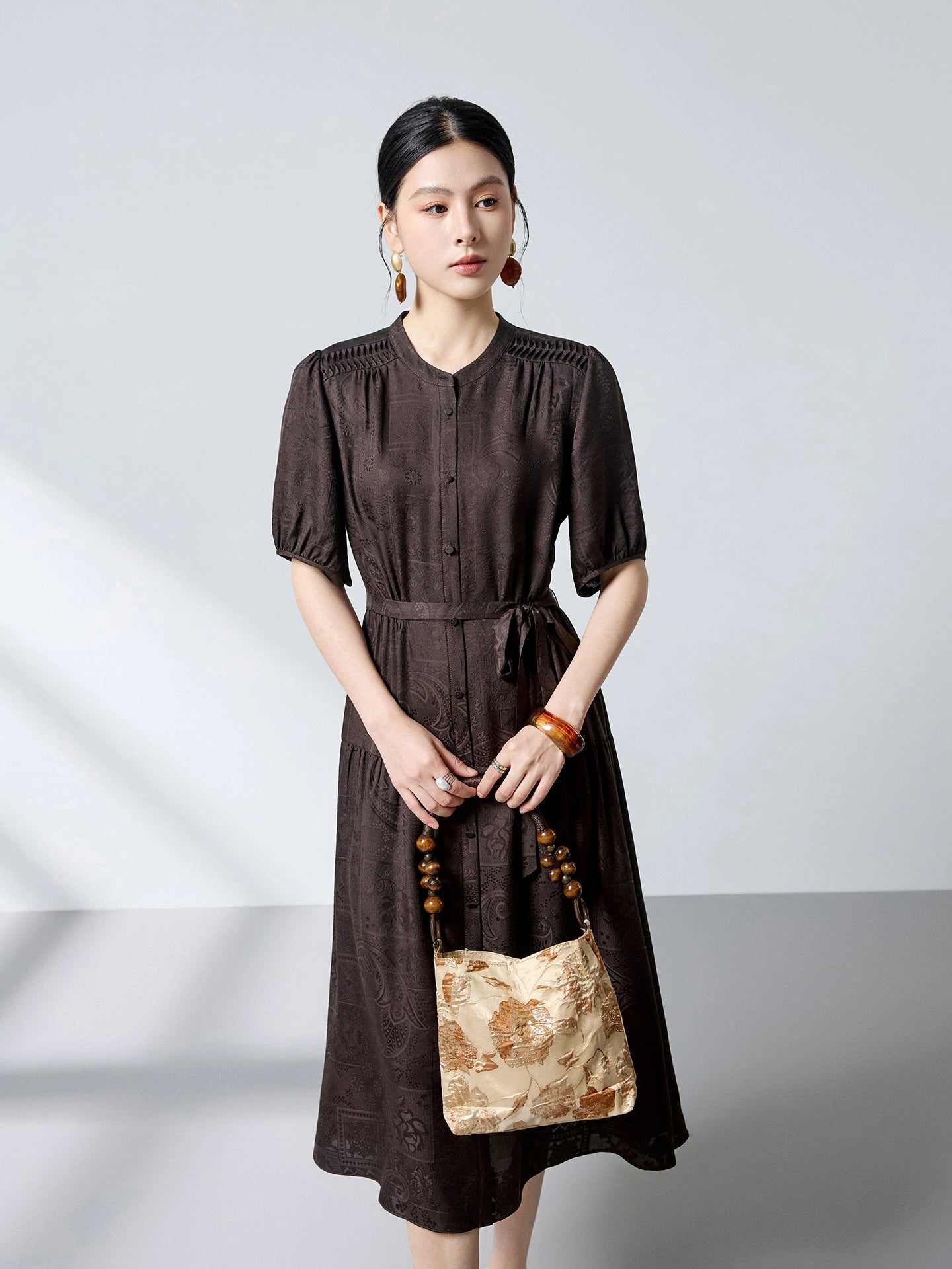 Full body shot of a brown silk jacquard dress with a button-down front and tie waist, styled with a beige handbag and beaded accessories.