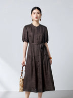 Model wearing a brown 100% silk jacquard dress with a round neckline, button-down front, and a tiered skirt, holding a beige handbag.