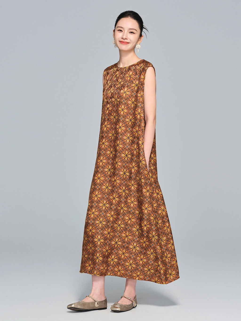 Model wearing a caramel brown 95%+ mulberry silk sleeveless dress with a round neckline and intricate geometric floral print, standing with hands in pockets.