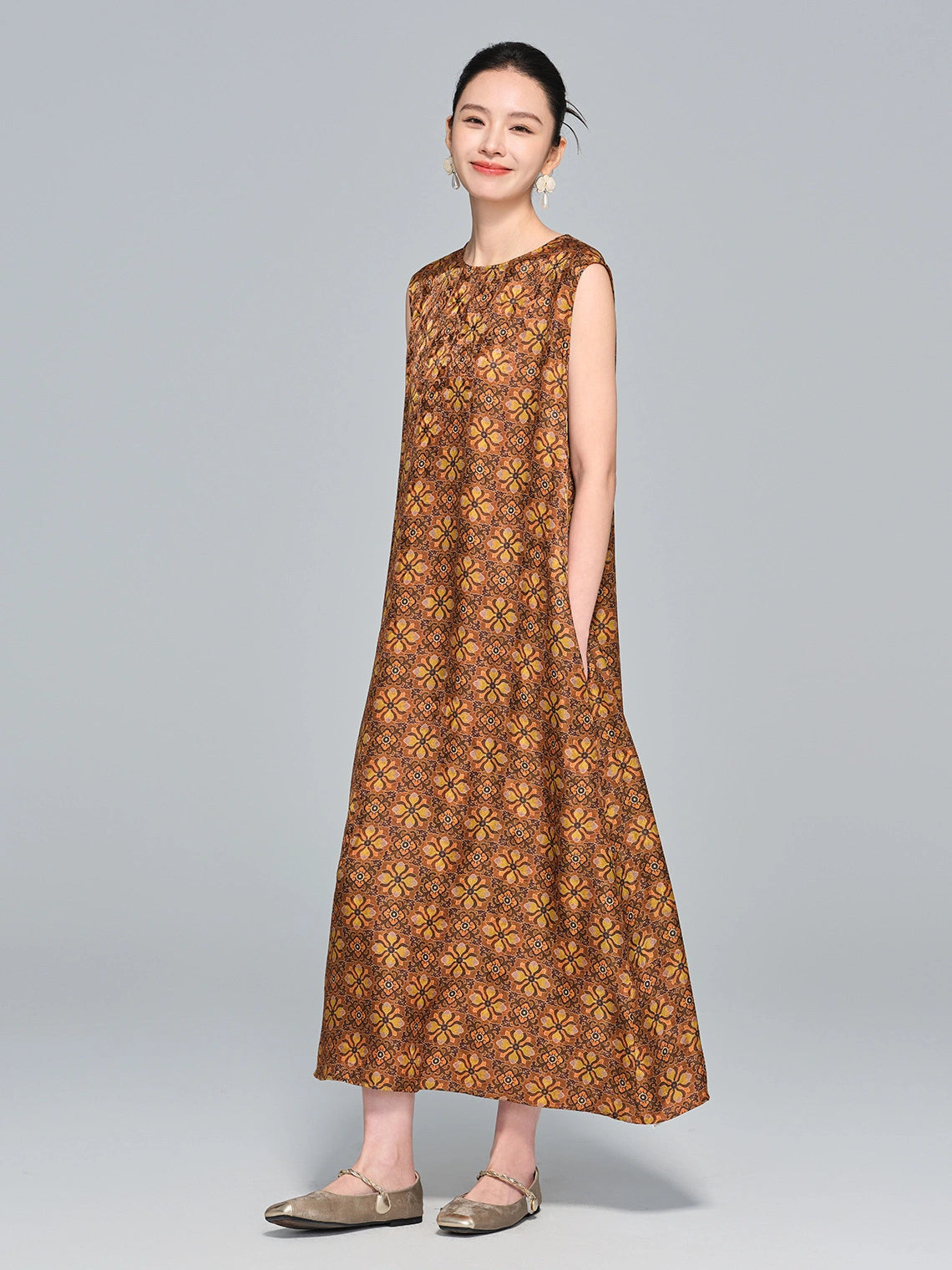 Model wearing a caramel brown 95%+ mulberry silk sleeveless dress with a round neckline and intricate geometric floral print, standing with hands in pockets.