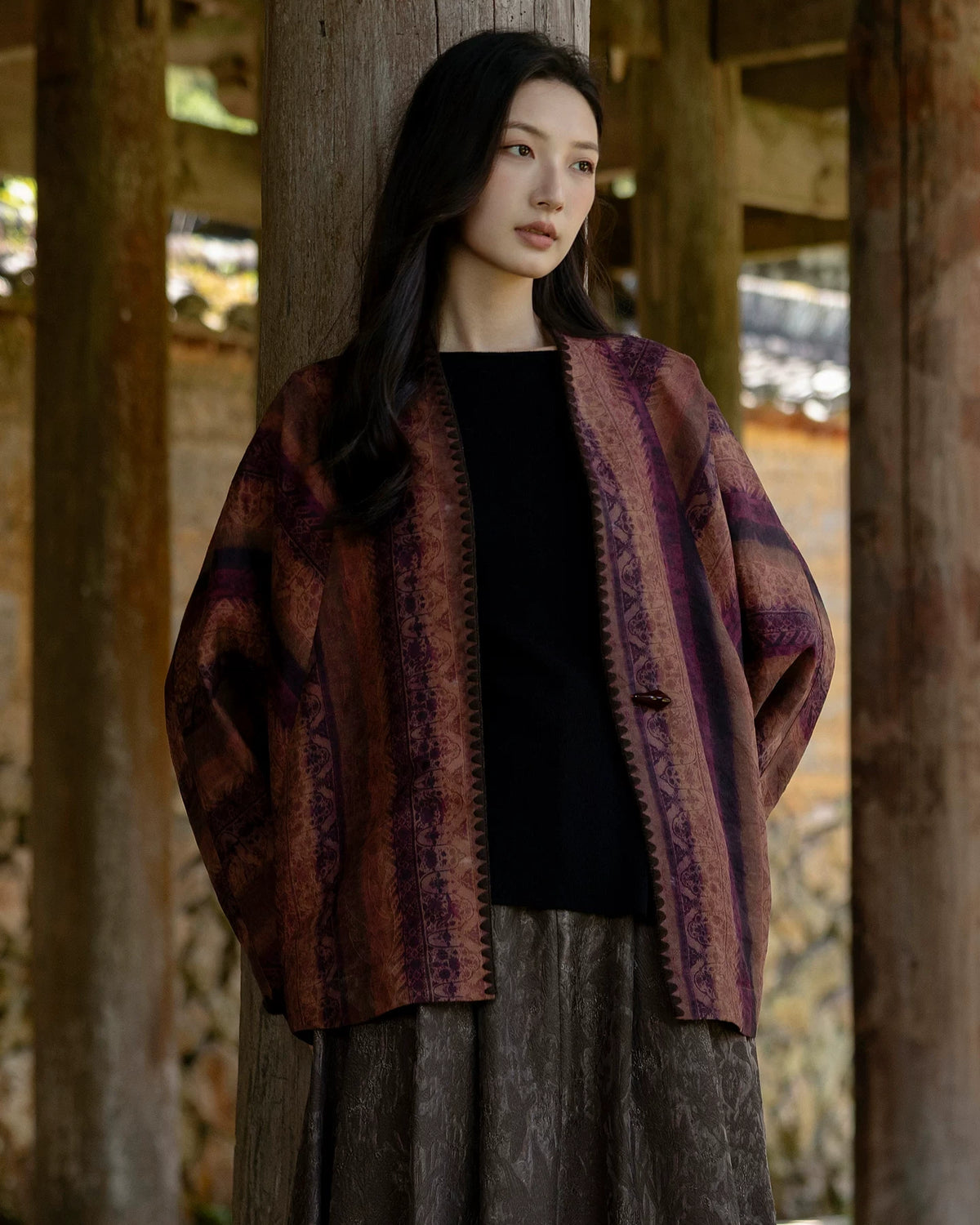 Model wearing the rust and plum patterned Canglan jacket open over a black top beside a wooden column.
