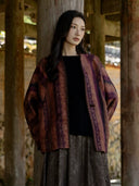 Model wearing the rust and plum patterned Canglan jacket open over a black top beside a wooden column.