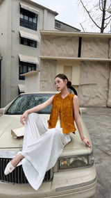 Woman sitting on a vintage car hood wearing a caramel brown sleeveless Cheongsam vest with mandarin collar, paired with white wide-leg pants.