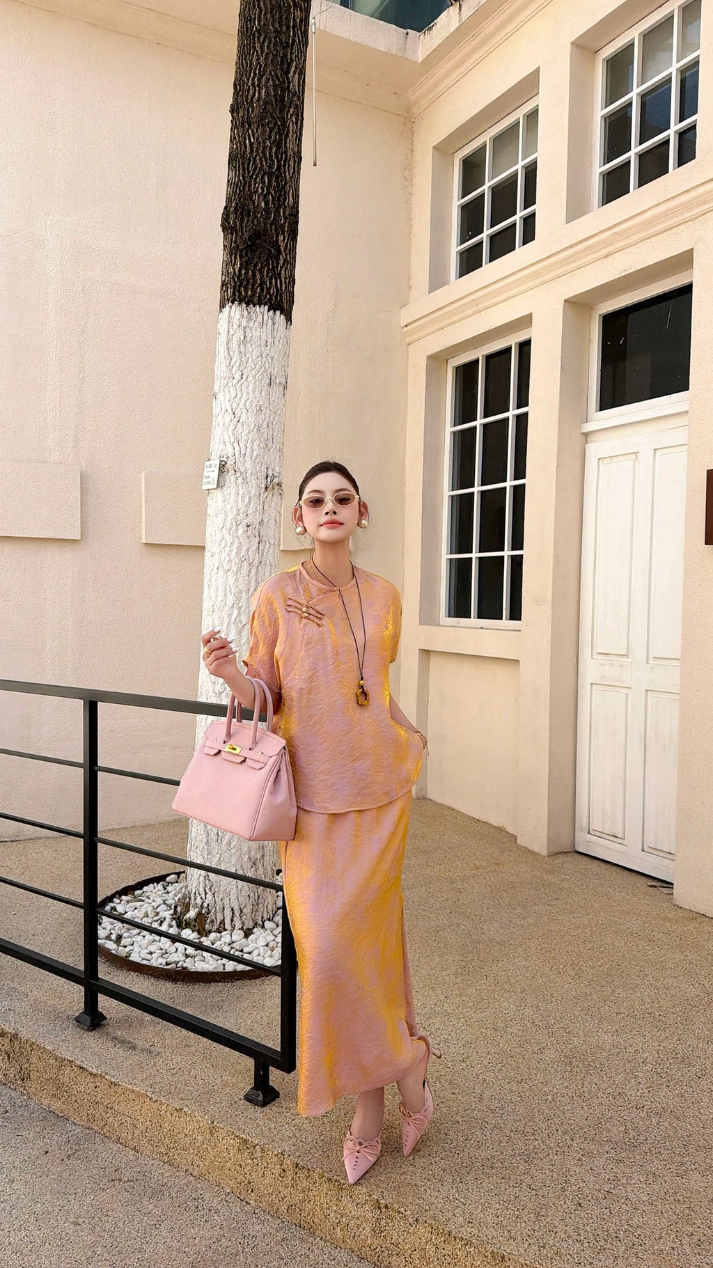Front view of a women's blush pink Qipao blouse and skirt set, featuring a mandarin collar and relaxed fit, styled with pink handbag and sunglasses.