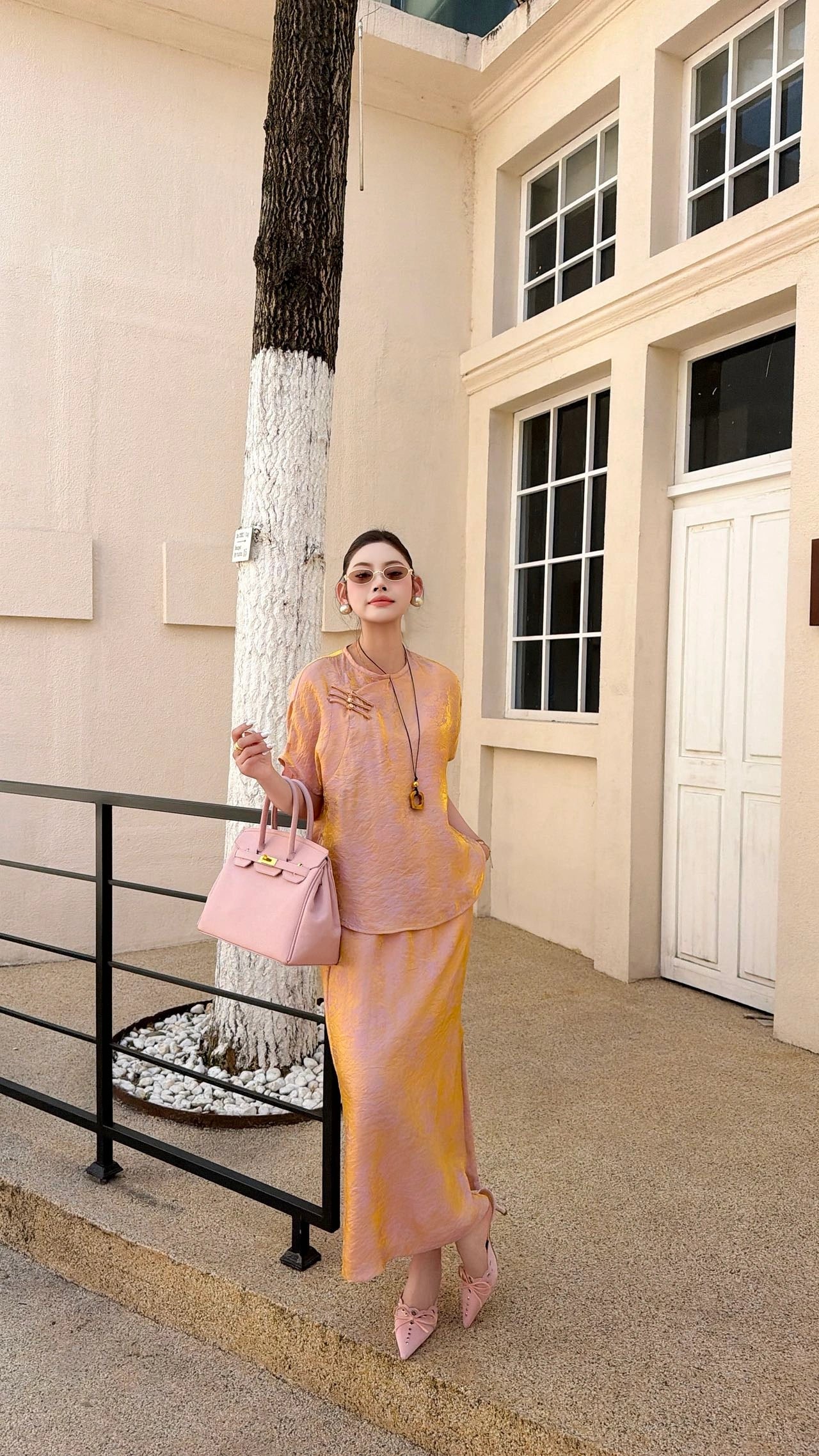 Front view of a women's blush pink Qipao blouse and skirt set, featuring a mandarin collar and relaxed fit, styled with pink handbag and sunglasses.