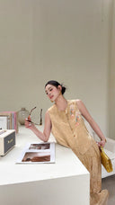 Woman leaning on a white counter wearing a champagne qipao vest set, holding sunglasses and a gold handbag.