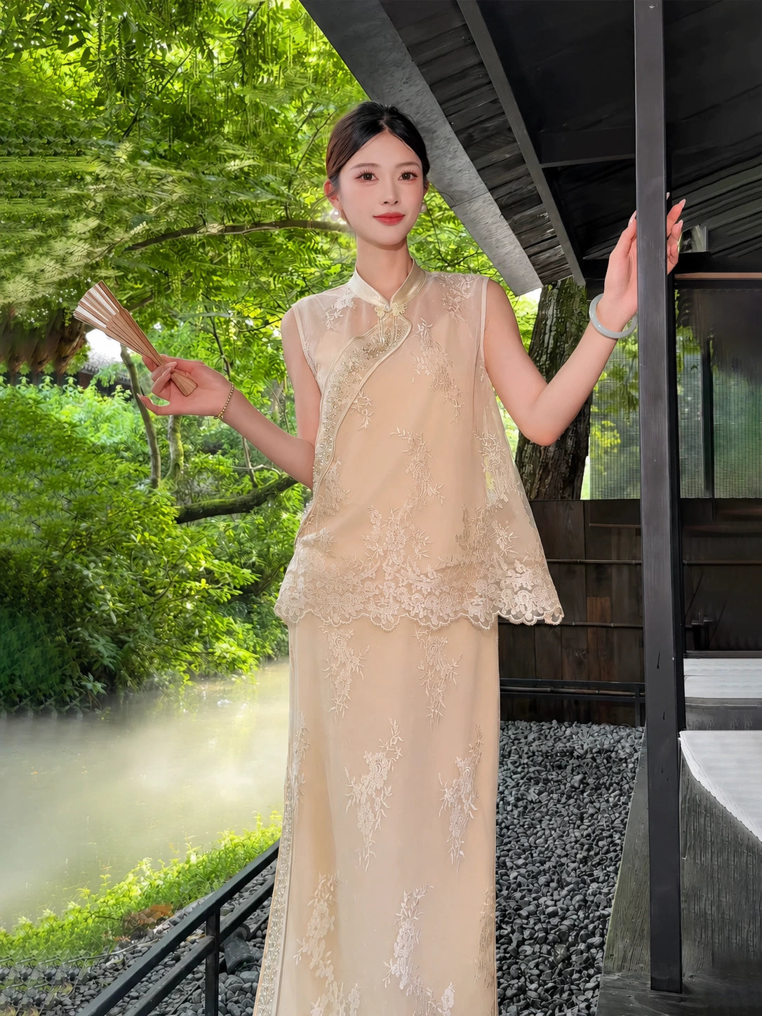Model wearing a champagne gold sleeveless Cheongsam vest with bead embroidery and lace hem, paired with a matching skirt, holding a fan in a garden setting.