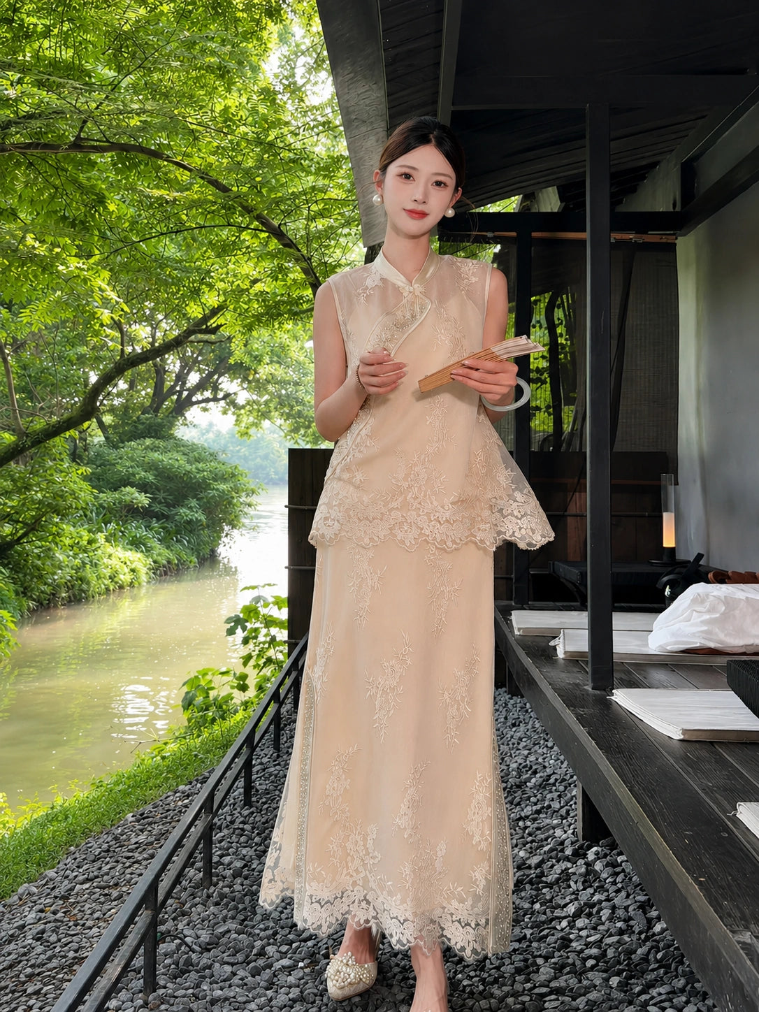 Model wearing a champagne gold Cheongsam vest with scalloped lace hem and matching embroidered skirt, holding a fan in a garden setting.