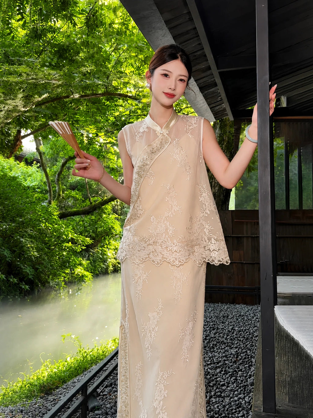 Full body shot of a champagne gold sleeveless vest and midi skirt set with bead embroidery and lace details, holding a folded fan.
