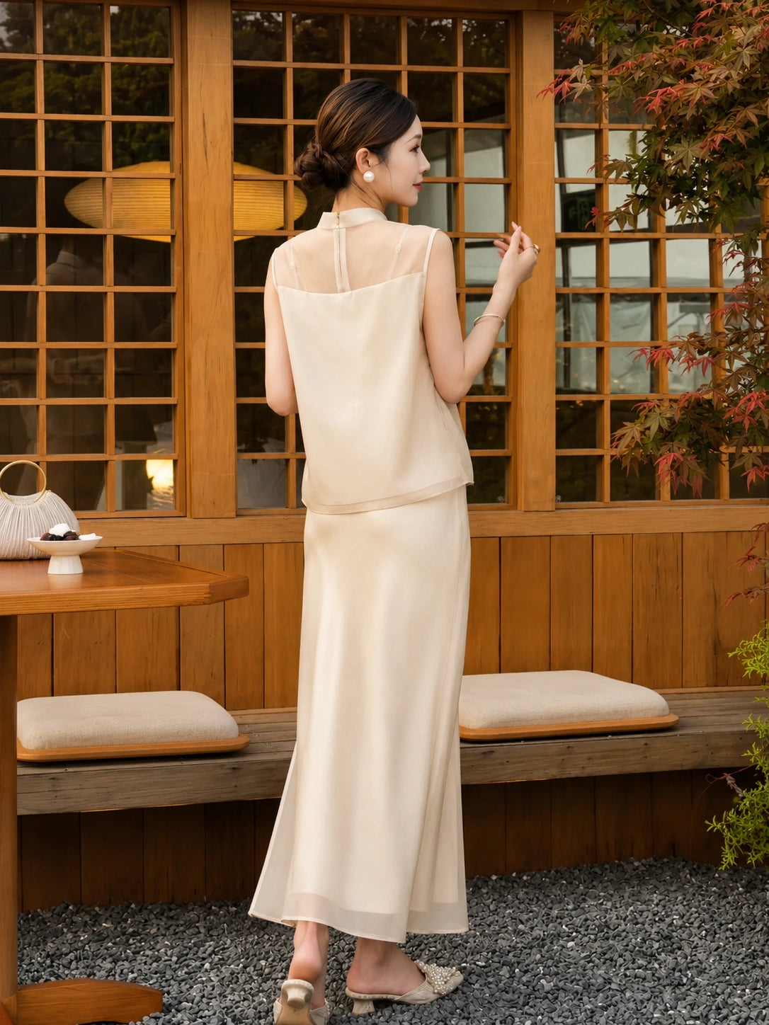Back view of a champagne Cheongsam set with a sheer panel and delicate floral appliqués, holding a fan, highlighting the smooth drape of the fabric.