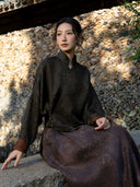 Seated model wearing the charcoal olive silk blouse, showing its stand collar, shaped hem and rust-brown turned-back cuffs.