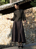 Sunlit front view of the charcoal olive blouse, showing its textured surface and contrasting turned-back cuffs.