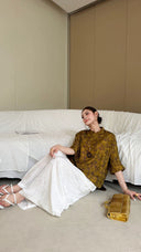 Sitting model in a metallic Qipao blouse with loose white pants, highlighting the 32% Lyocell fabric drape and comfortable Cheongsam top design.
