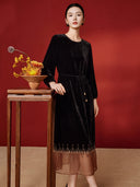 Model wearing the black velvet midi dress with a tie waist and copper-toned sheer hem beside autumn flowers