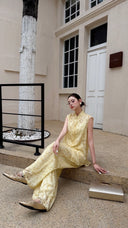 Woman sitting on stone steps wearing a cream yellow Cheongsam top and pants set, with a silver handbag on the ground.