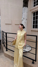 Woman wearing a cream yellow Cheongsam top with mandarin collar, paired with matching wide-leg trousers, holding a gold bag.