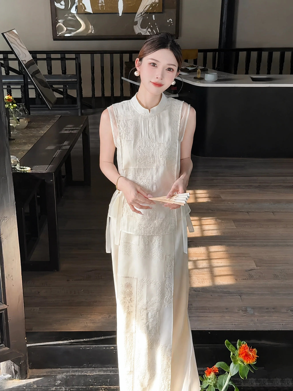 Model wearing a cream white sleeveless Cheongsam vest with side ties and a matching embroidered midi skirt, holding a paper fan.