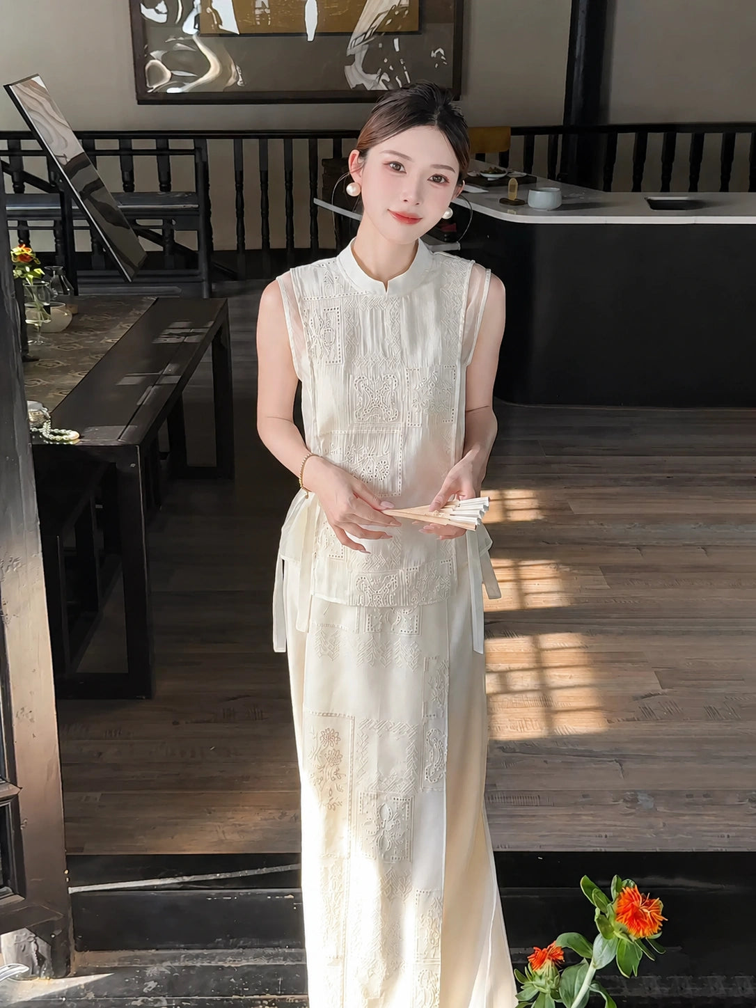 Model wearing a cream white sleeveless Cheongsam vest with side ties and a matching embroidered midi skirt, holding a paper fan.